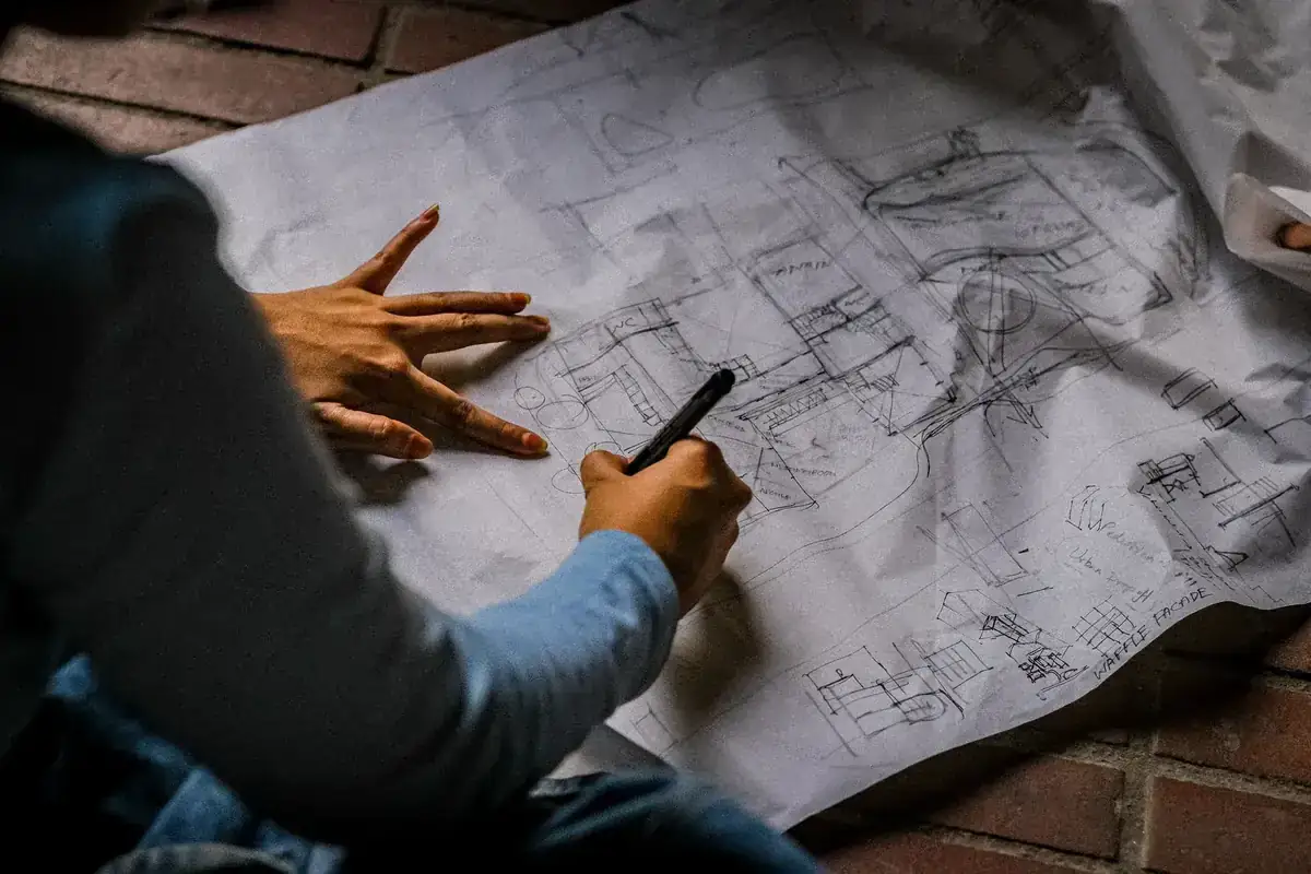 Person sketching architectural design on blueprint paper with pen on brick floor