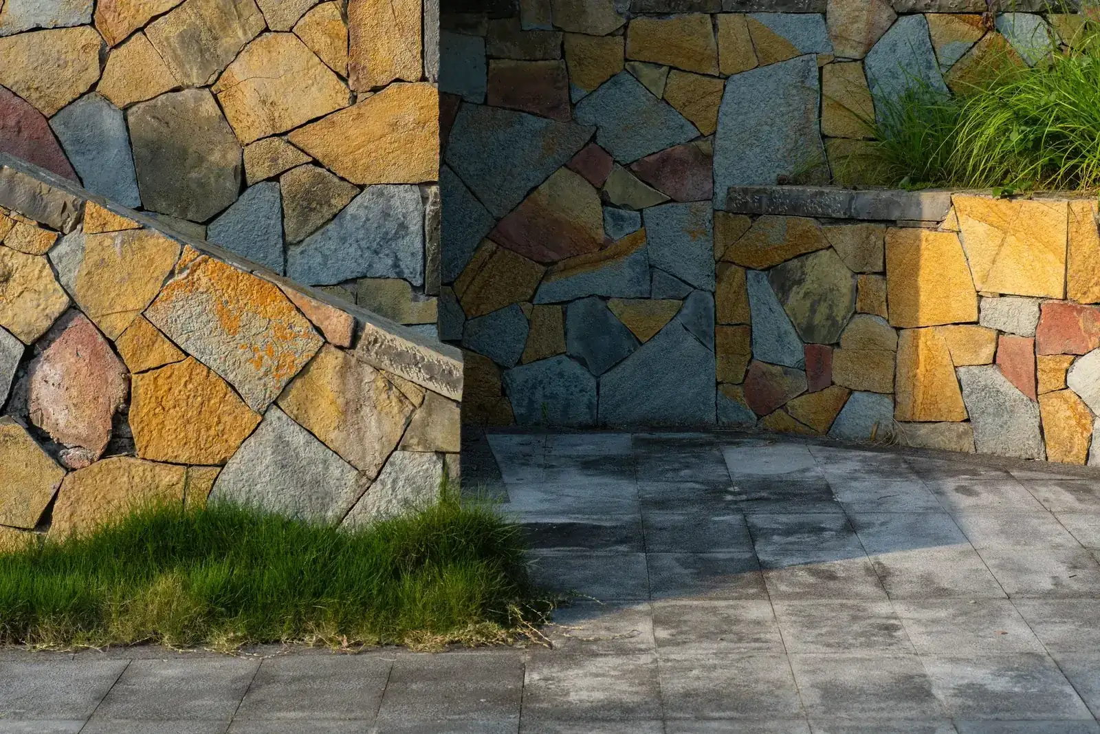 Close-up of a colorful stone wall with paved ground and lush grass in sunlight