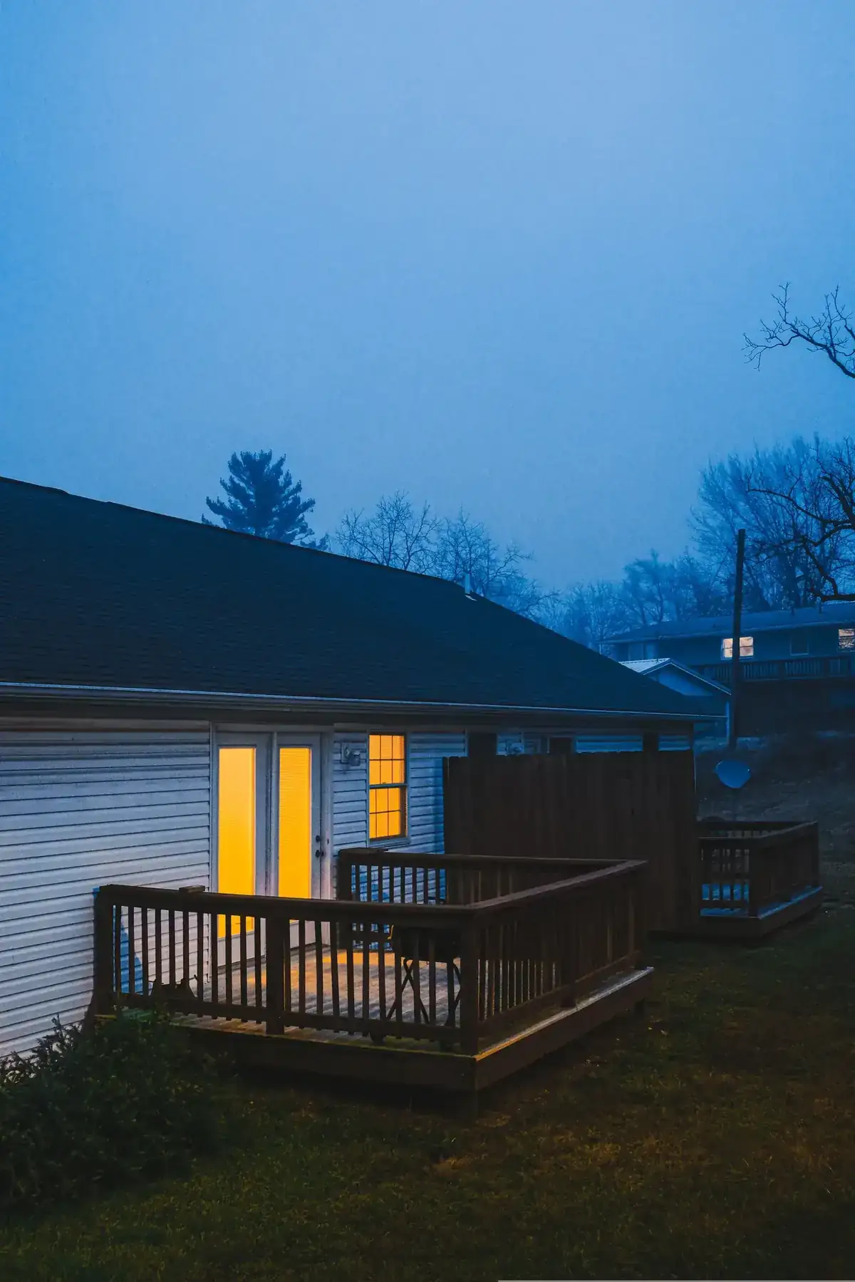 A suburban house with a warmly lit porch on a misty evening outdoors