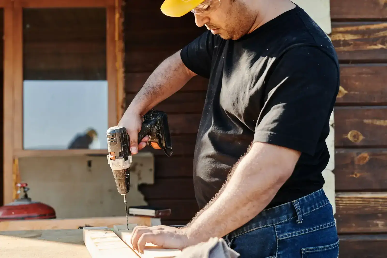 A construction worker wearing a helmet uses a power drill on a wooden plank at an outdoor construction site